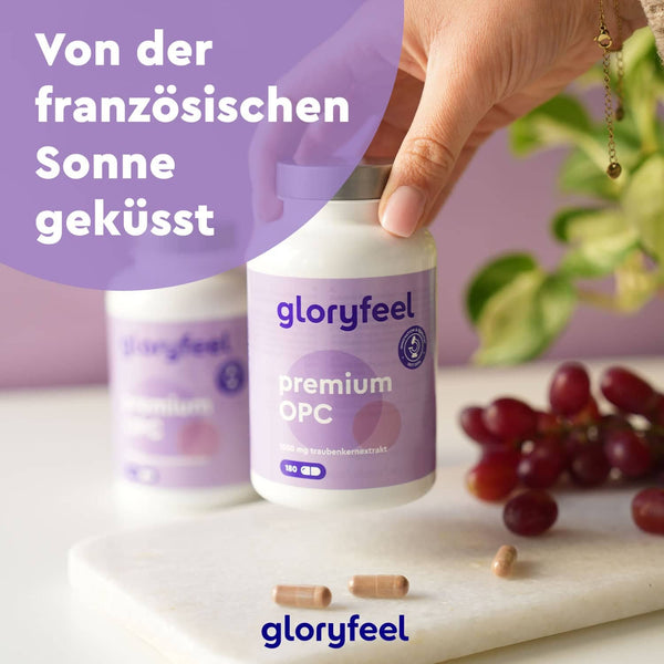 Eine Hand hält eine Flasche Premium OPC 180 Kapseln der gloryfeel GmbH neben Weintrauben, einem Schneidebrett und Kapseln; das Bild zeigt einen deutschen Text.