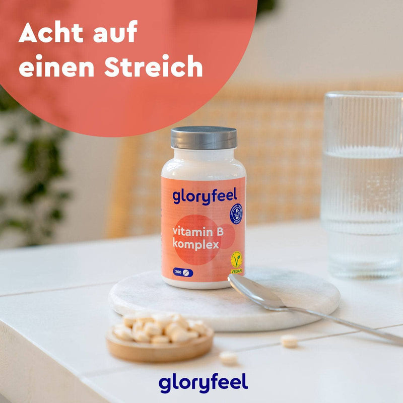 Eine Flasche Vitamin B Komplex der gloryfeel GmbH steht auf dem Tisch, umgeben von verstreuten Pillen, einem Glas Wasser und einem Löffel. Der deutsche Text oben hebt die Vorteile für Wohlbefinden und Stoffwechsel hervor.