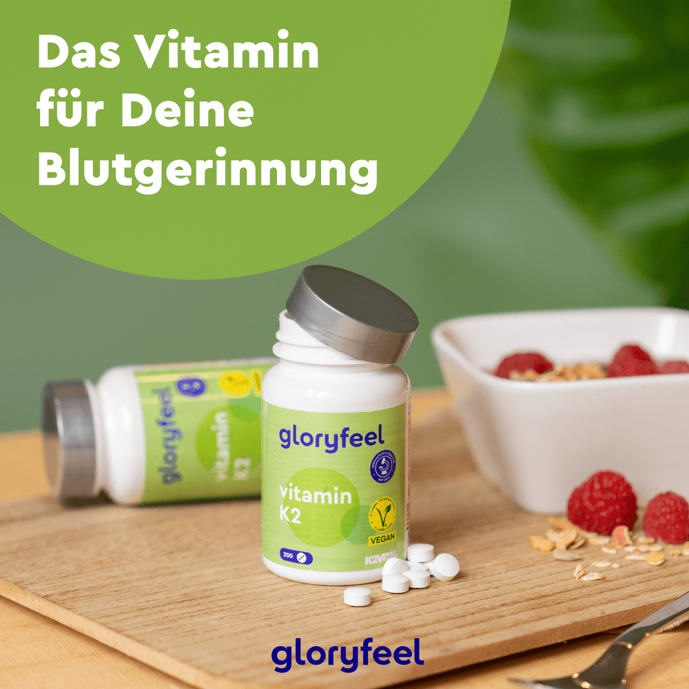 Zwei Flaschen Vitamin K2 Tabletten der Gloryfeel GmbH, ein Nahrungsergänzungsmittel, stehen auf einer Holzoberfläche neben Müsli mit Himbeeren. Text lautet: „Das Vitamin für Deine Blutgerinnung.