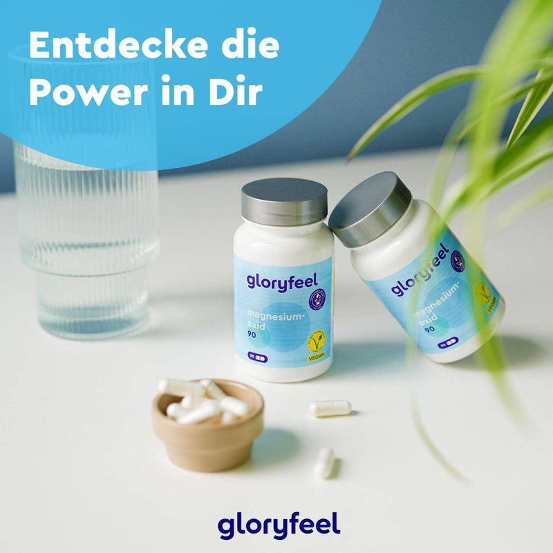 Zwei Flaschen Magnesiumoxid 90 Kapseln der gloryfeel GmbH stehen auf einem Tisch mit verstreuten Kapseln, einer Pflanze und einem Glas. Der deutsche Text oben lautet: „Entdecke die Power in Dir.“.