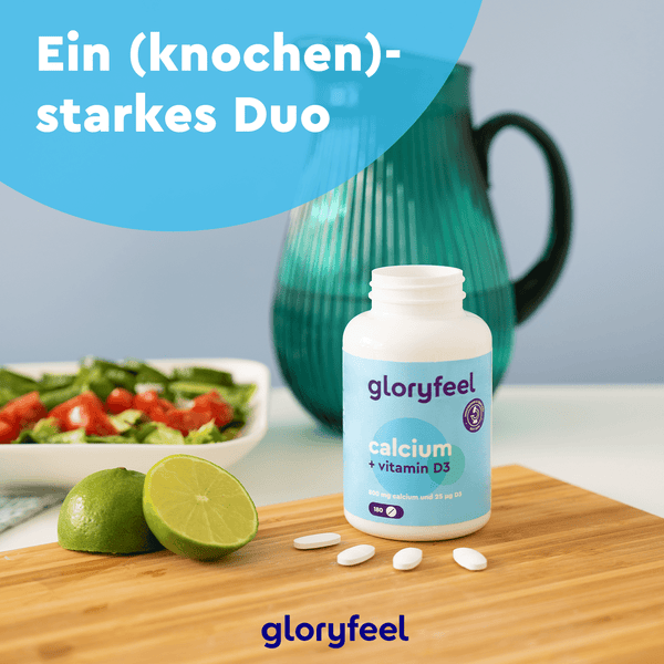 Eine Flasche „Calcium + Vitamin D3 Tabletten“ der gloryfeel GmbH steht auf einem Holzbrett neben einer Limettenscheibe, im Hintergrund sind eine Salatschüssel und ein türkisfarbener Krug zu sehen.
