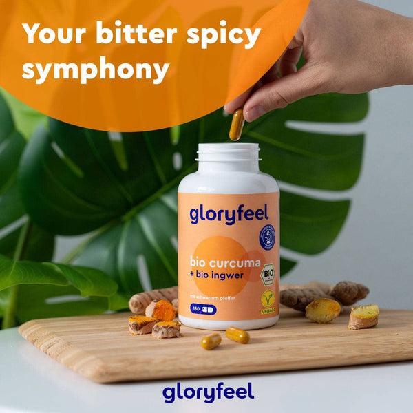 Eine Hand hält eine Bio Curcuma + Bio Ingwer Kapseln über einer geöffneten Flasche mit der Aufschrift "gloryfeel GmbH". Im Hintergrund sind frische Kurkumawurzeln und grüne Blätter zu sehen.