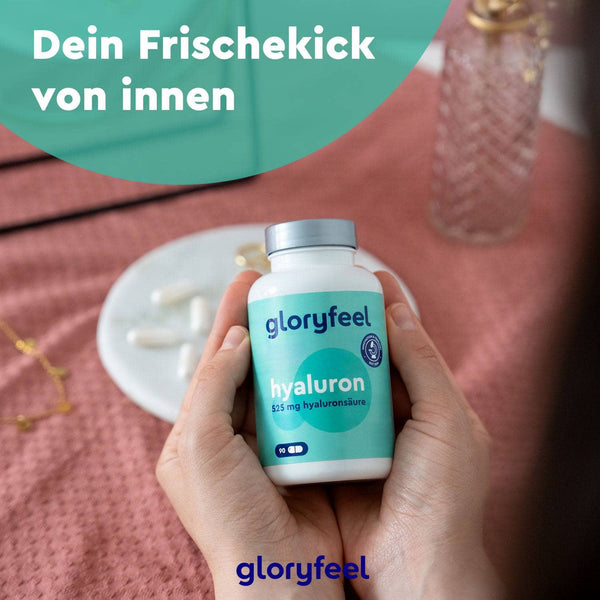 Hände halten eine Flasche Hyaluron Kapseln Intensiv der gloryfeel GmbH. Der deutsche Text lautet: „Dein Frischekick von innen“. Im Hintergrund sind ein Glas, einige Kapseln und ein rosa Tuch zu sehen.