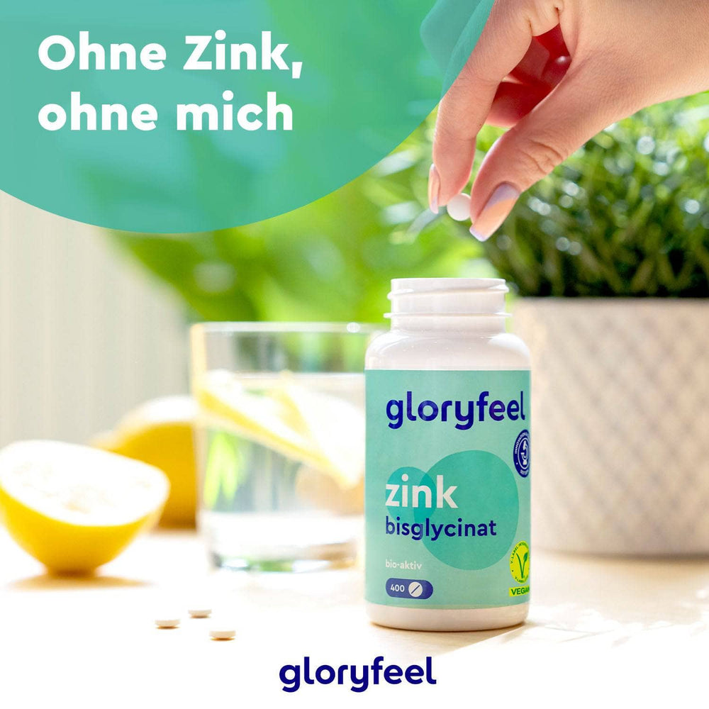 Eine Hand hält eine Flasche mit der Aufschrift „gloryfeel GmbH Zinkbisglycinat Tabletten“ über einem Tisch mit Zitronen und einem Glas Wasser. Der deutsche Text lautet: „Ohne Zink, ohne mich.“ Dieses Nahrungsergänzungsmittel unterstreicht das essentielle Wohlbefinden.