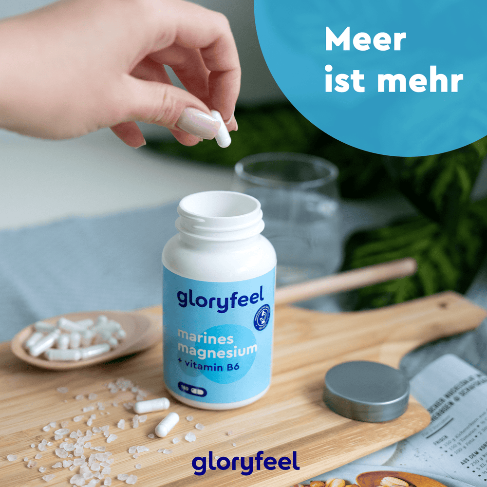 Eine Hand greift nach einer Kapsel aus einer Flasche mit der Aufschrift „Marines Magnesium Kapseln“ der gloryfeel GmbH auf einem Holzbrett, wodurch ihre Rolle bei der Unterstützung der Muskelfunktion hervorgehoben wird. In der Nähe sind weitere Magnesiumkapseln verstreut.