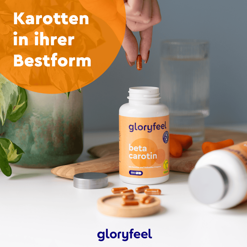Eine Person hält eine Nahrungsergänzungskapsel über einer offenen Flasche mit der Aufschrift „gloryfeel GmbH Beta-Carotin Kapseln“ auf einem Tisch mit Wasser und einer Pflanze. Der deutsche Text lautet: „Karotten in ihrer Bestform.“.