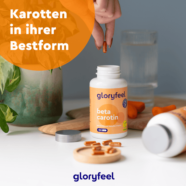 Eine Person hält eine Nahrungsergänzungskapsel über einer offenen Flasche mit der Aufschrift „gloryfeel GmbH Beta-Carotin Kapseln“ auf einem Tisch mit Wasser und einer Pflanze. Der deutsche Text lautet: „Karotten in ihrer Bestform.“.