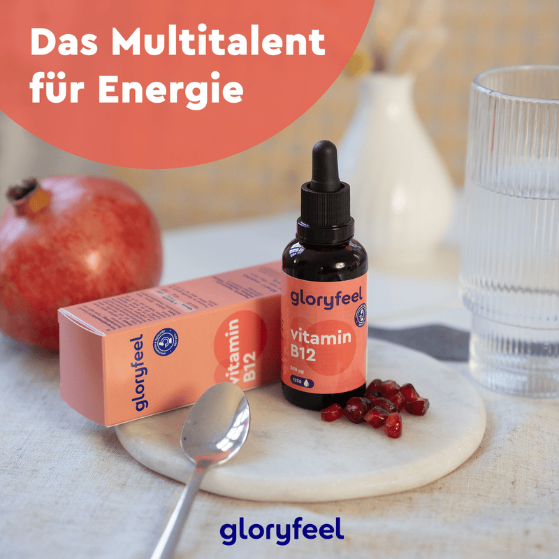 Eine Vitamin-B12-Tropfenflasche der gloryfeel GmbH steht mit einer Pipette auf einem Tisch, daneben eine Schachtel, verstreute Granatapfelkerne, ein Löffel und ein Glas. Ein orangefarbener Kreis zeigt einen deutschen Text zum Thema Energie.