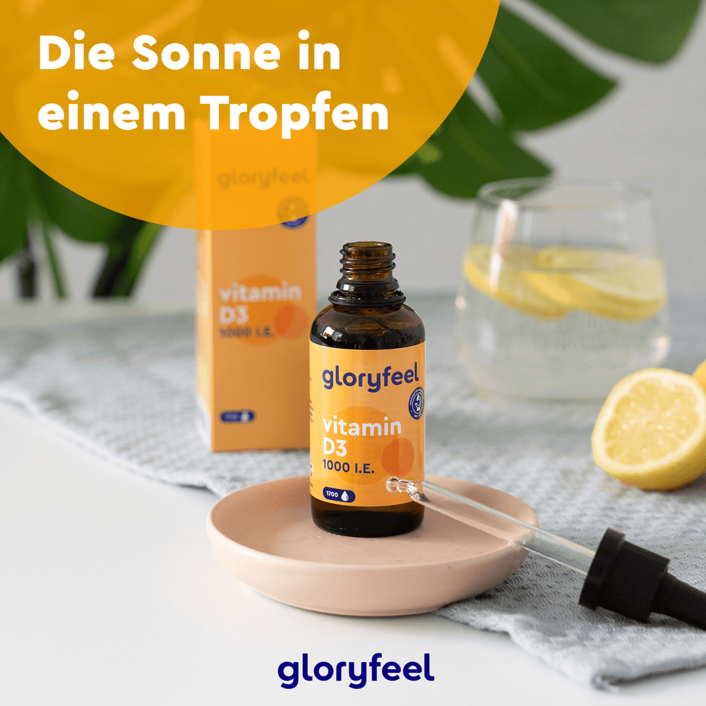 Eine Flasche Vitamin D3 Tropfen 1000 I.E. der gloryfeel GmbH steht mit einer Pipette auf einem kleinen Teller, umgeben von einem Glas Zitronenwasser und Zitronenscheiben auf dem Tisch. Der deutsche Text lautet: „Die Sonne in einem Tropfen.“.