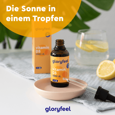 Eine Flasche Vitamin D3 Tropfen 1000 I.E. der gloryfeel GmbH steht mit einer Pipette auf einem kleinen Teller, umgeben von einem Glas Zitronenwasser und Zitronenscheiben auf dem Tisch. Der deutsche Text lautet: „Die Sonne in einem Tropfen.“.