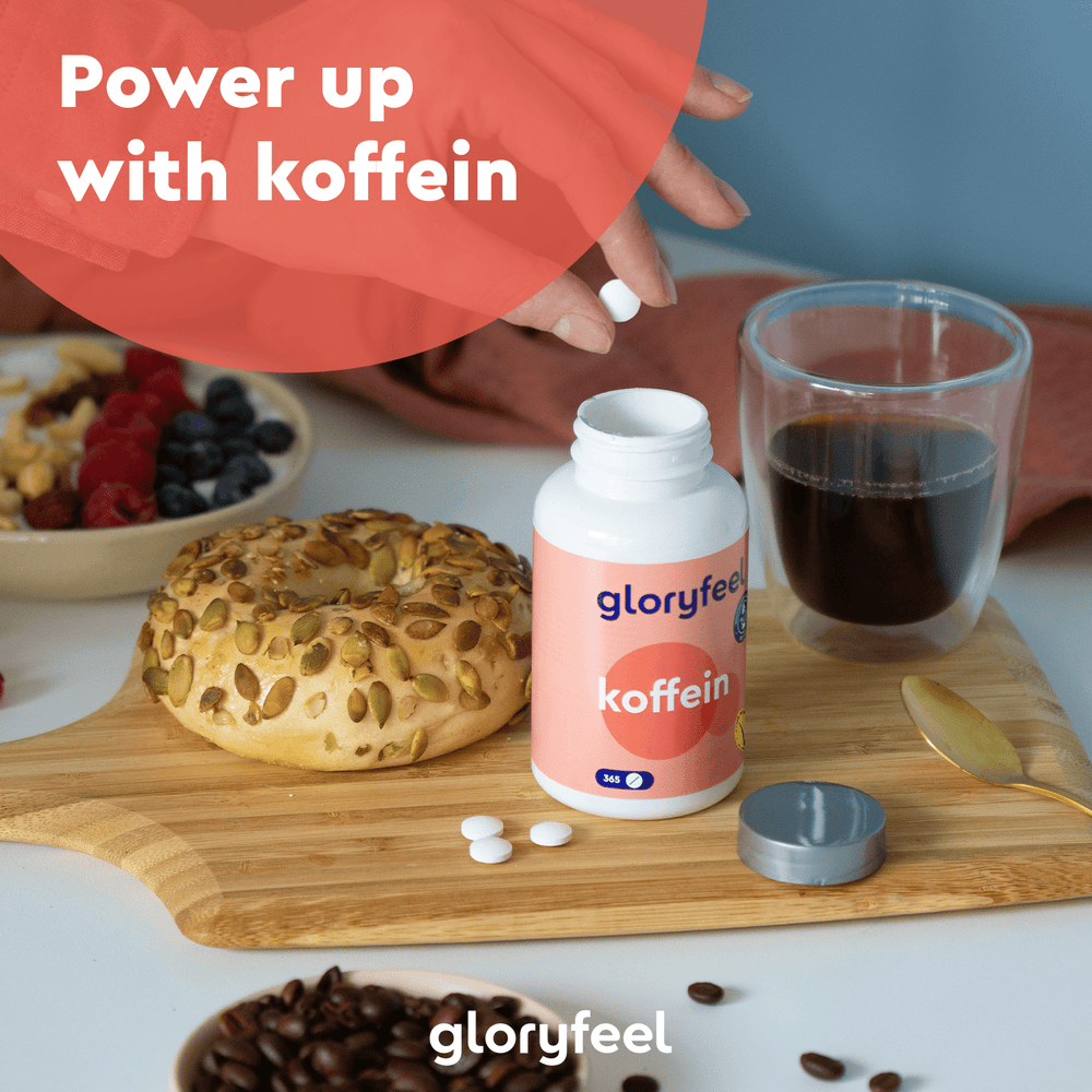 Auf einem Tisch stehen eine Tasse Kaffee, ein Bagel mit Kürbiskernen und eine Flasche Koffeintabletten der gloryfeel GmbH. Eine Hand hält eine Pille. Kaffeebohnen und Beeren liegen verstreut herum.