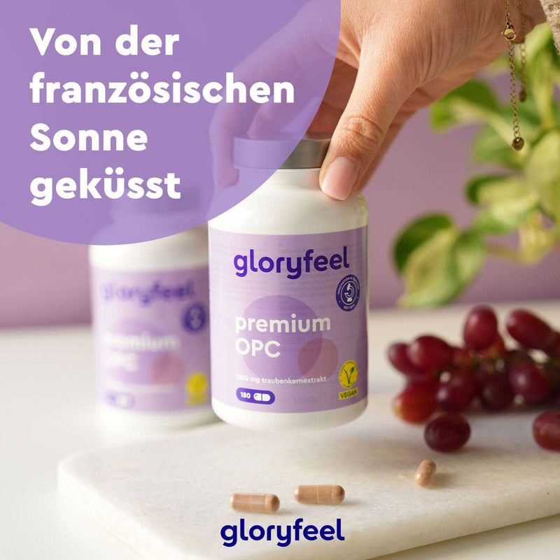Zwei Flaschen Premium OPC 180 Kapseln von Gloryfeel auf einem Tisch mit Kapseln und Weintrauben, eine davon von einer Hand gehalten. Text auf Deutsch: „Von der französischen Sonne geküsst.