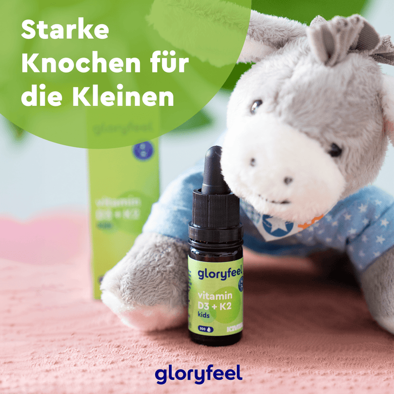 Eine Gloryfeel Vitamin D3 K2 Kids Tropfenflasche steht neben einem Plüschesel auf rosa. Der deutsche Text „Starke Knochen für die Kleinen“ betont die Vorteile von Vitamin K2 (MK-7) für die Knochenstärke von Kindern. Marke: gloryfeel GmbH.