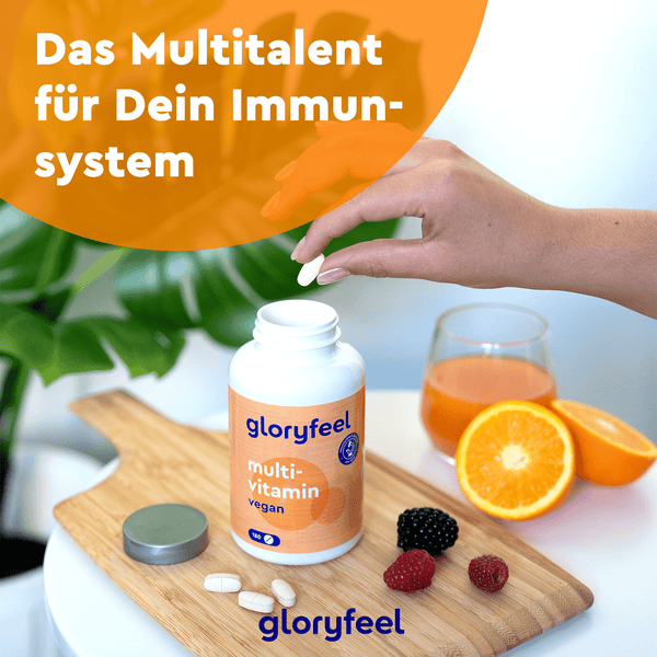 Eine Hand greift nach einer Kapsel aus einer Flasche veganer Multivitamintabletten der gloryfeel GmbH auf einem Tisch mit Früchten, Saft und Natur. Der Bildtext betont die Vorteile natürlicher Vitamine und die Unterstützung des Immunsystems für Menschen, die sich vegan ernähren.