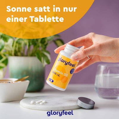 Eine Hand hält eine Flasche Vitamin D3 Tabletten 4000 I.E. der gloryfeel GmbH neben einer Keramikschale und einem Glas Wasser. Im Hintergrund ist eine Pflanze zu sehen. Der Text lautet: „Sonne satt in nur einer Tablette.“.