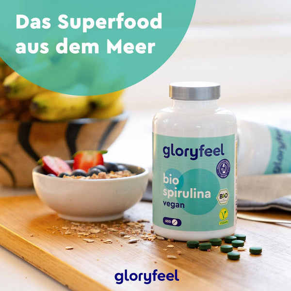 Eine Flasche Bio Spirulina Tabletten der gloryfeel GmbH steht elegant auf dem Tisch neben einer frischen Schüssel Müsli, frischen Früchten und einer Bananenstaude. Der Text „Das Superfood aus dem Meer“ hebt die reichhaltigen Vorteile dieses Bio-Nahrungsergänzungsmittels hervor.