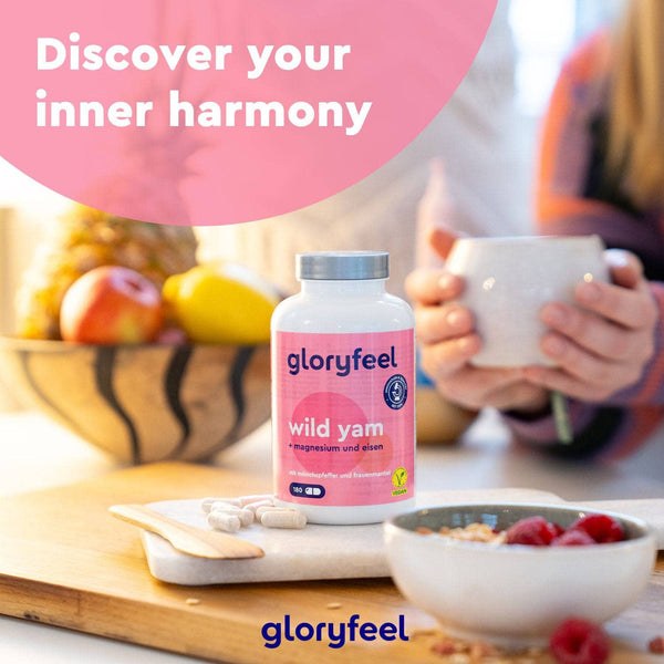 Eine Flasche Wild Yam Kapseln der gloryfeel GmbH steht auf einem Tisch mit Früchten, Pillen und einer Person, die eine Tasse hält. In einer rosa Sprechblase steht: „Entdecke deine innere Harmonie.“.